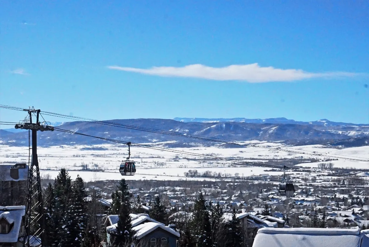 This expansive view captures the iconic gondola lift soaring over snow-covered mountains, with pine trees and a clear blue sky setting an adventurous, wintry mood. The image embodies the excitement and scenic beauty of Steamboat’s ski terrain.