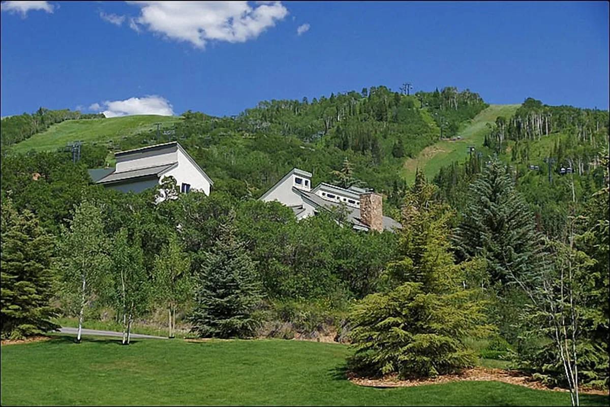 The image displays the exterior of the PowderGlades Chalet nestled among lush green trees, with rolling hills and mountain scenery in the background. The house is surrounded by nature, suggesting direct access to hiking and biking trails, with ample space for outdoor activities and relaxation.