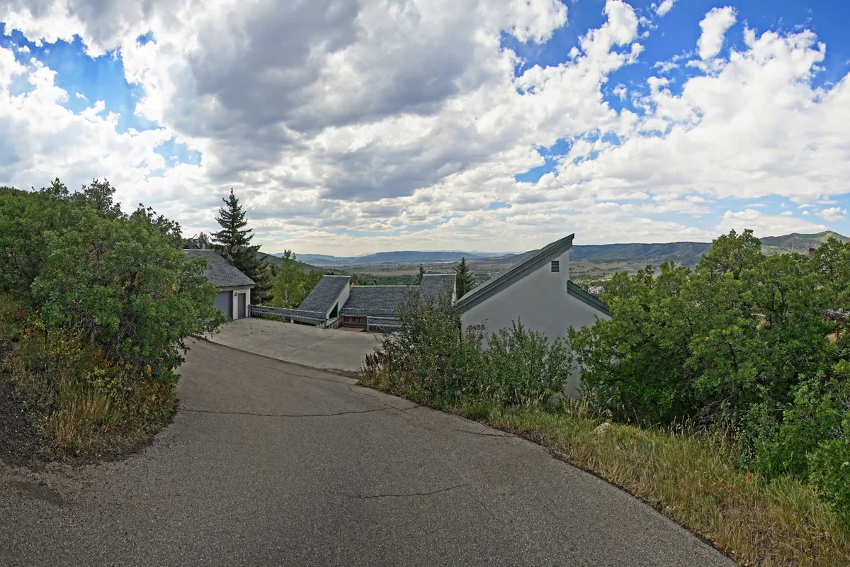 An exterior view of PowderGlades Chalet showing a winding driveway surrounded by lush greenery, with distant mountains and a cloudy sky in the background. The peaceful setting emphasizes the home's connection to nature and the sense of retreat it offers.