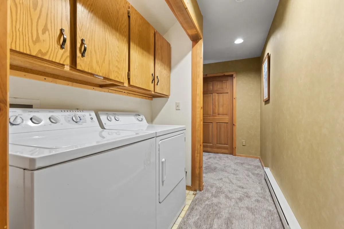 The laundry room features a modern washing machine and dryer, complemented by wooden cabinets for storage, soft plush carpeting, and framed artwork. The space is functional while maintaining a homey, comfortable vibe, ideal for longer stays or active families.