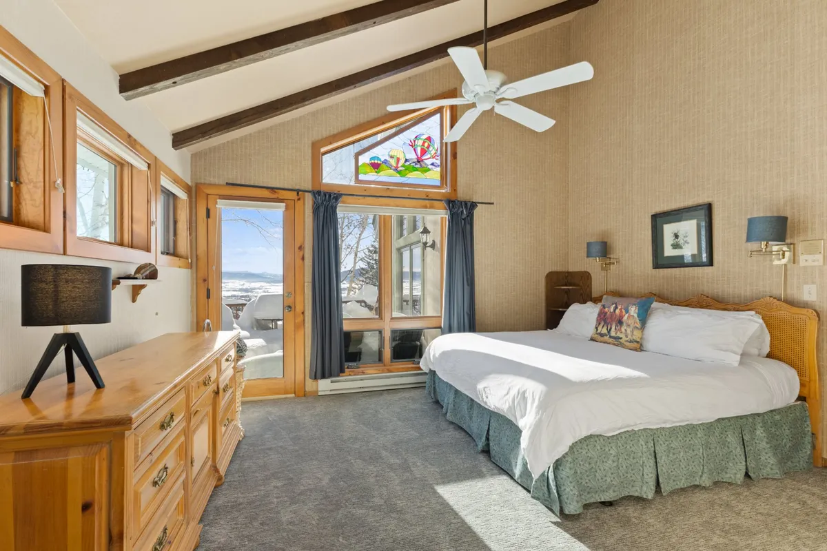 A spacious master bedroom featuring a large bed with plush linens, a beautiful stained glass window letting in natural light, a wooden dresser, and a ceiling fan. The calm and tranquil atmosphere makes it ideal for relaxation and enjoying the view outside.