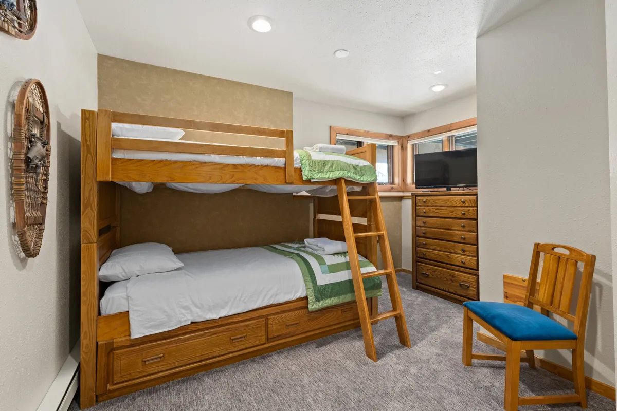 A charming bunk room with a wooden bunk bed, a dresser, a wooden chair, and a window bringing in natural light. The room is inviting, perfect for children or friends, and evokes the spirit of cozy sleepovers and shared memories.
