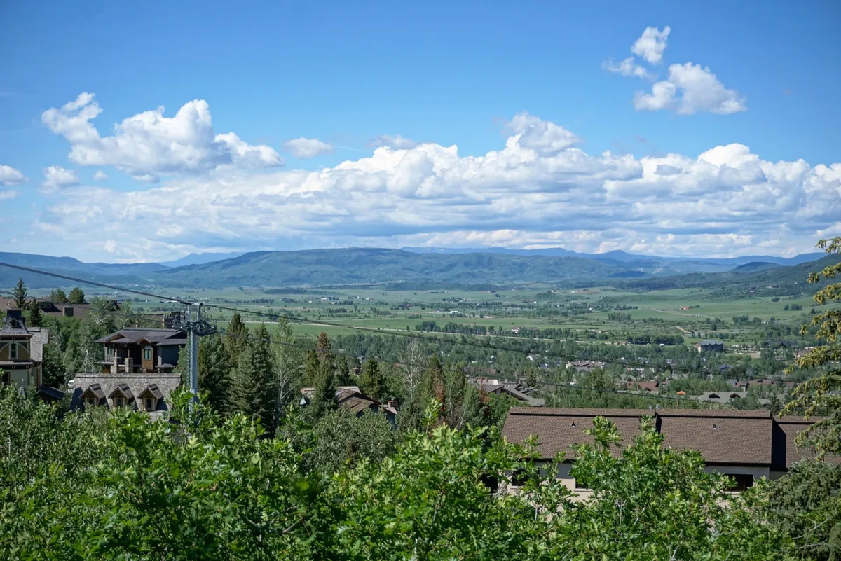 A sweeping view of the mountain landscape and valley, with cloud formations and homes in the foreground, emphasizing the natural beauty of the surrounding area. The setting sun casts a soft light, creating a serene and inviting atmosphere.