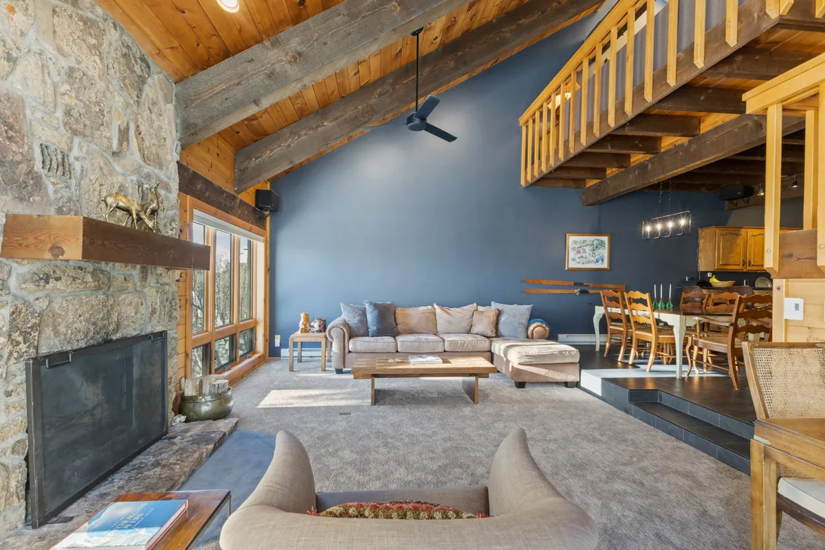 This inviting interior view highlights a cozy living room centered around a prominent stone fireplace, with plush sofas and exposed wood beams above. The adjoining dining area is visible, and the overall space is flooded with natural light, creating a warm and communal ambiance.
