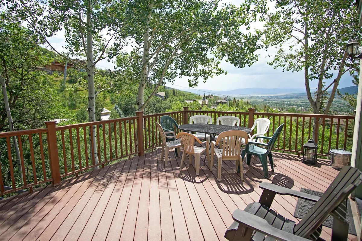 This exterior image centers on a round dining table with colorful chairs set on a wooden deck framed by trees. The mountain view in the background hints at sunrise coffees and starlit gatherings, underscoring the section’s atmosphere of relaxed outdoor dining and natural beauty.