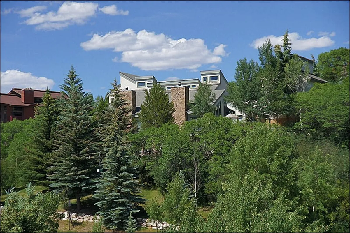 The image showcases the modern, inviting exterior of a single-family chalet surrounded by lush greenery and set against a brilliant blue sky dotted with fluffy clouds. The tranquil scene highlights the architectural charm of the home and the peaceful, natural beauty of Steamboat Springs, with distant mountain views hinting at nearby slopes.