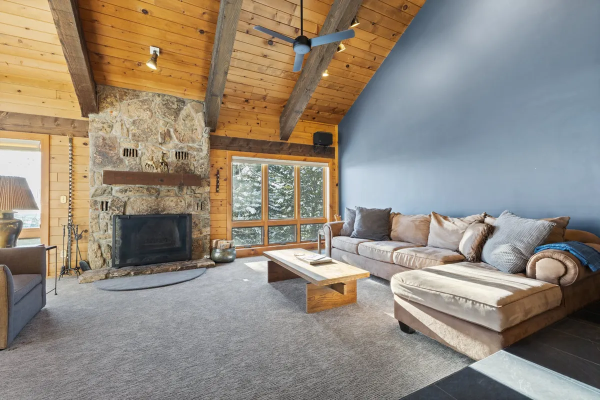 A living room anchored by a stone fireplace, large windows with views of nature, a comfortable sectional sofa, and a wooden coffee table. Natural light pours in, creating a warm and inviting place to unwind after a day outdoors.