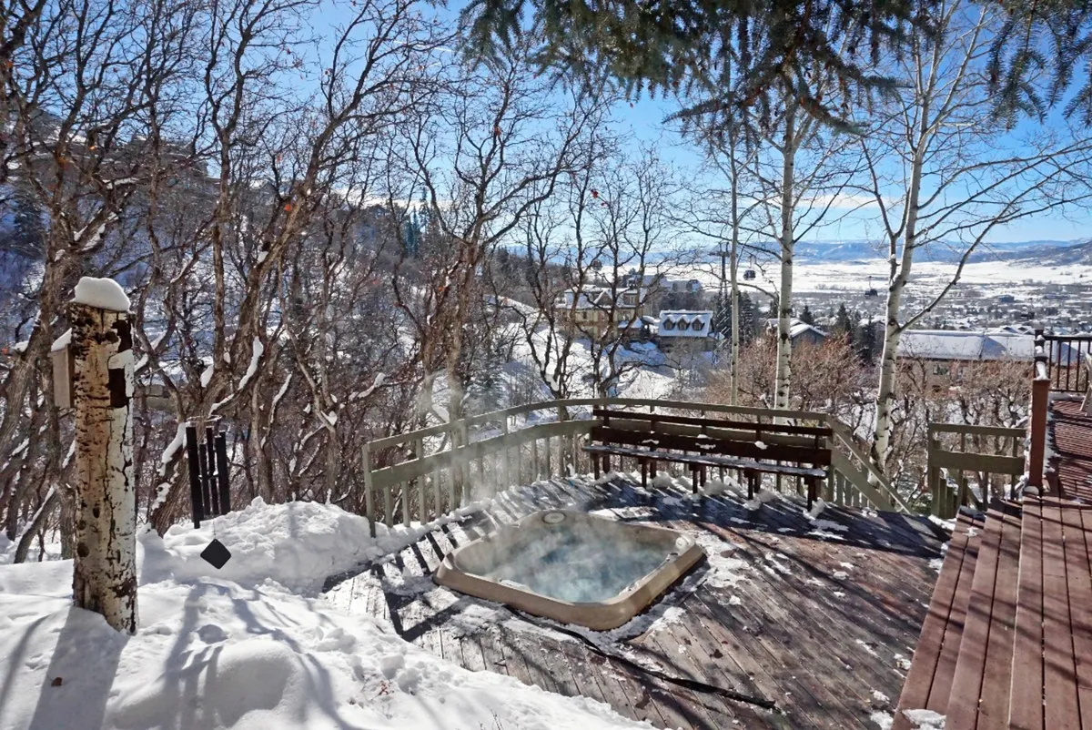 An inviting wooden deck with a hot tub overlooks a backdrop of trees and mountain views. The scene conveys tranquility, relaxation, and a perfect spot to unwind after a day of adventure, with potential for sunset or stargazing experiences.