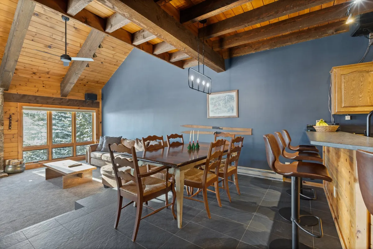 This angle offers a seamless view of the dining table, bar seating, large windows framing valley views, and rustic wooden ceiling beams. The layout fosters easy flow and togetherness, with sunlight streaming through the windows.