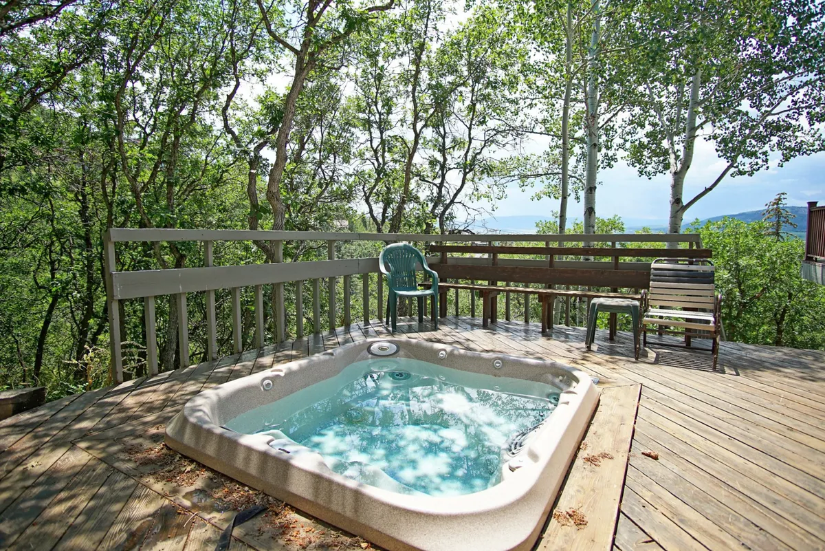 A tranquil corner of the deck featuring a hot tub and built-in bench seating, surrounded by evergreens and aspens. The trees provide privacy while inviting the eye outward toward glimpses of nature beyond.