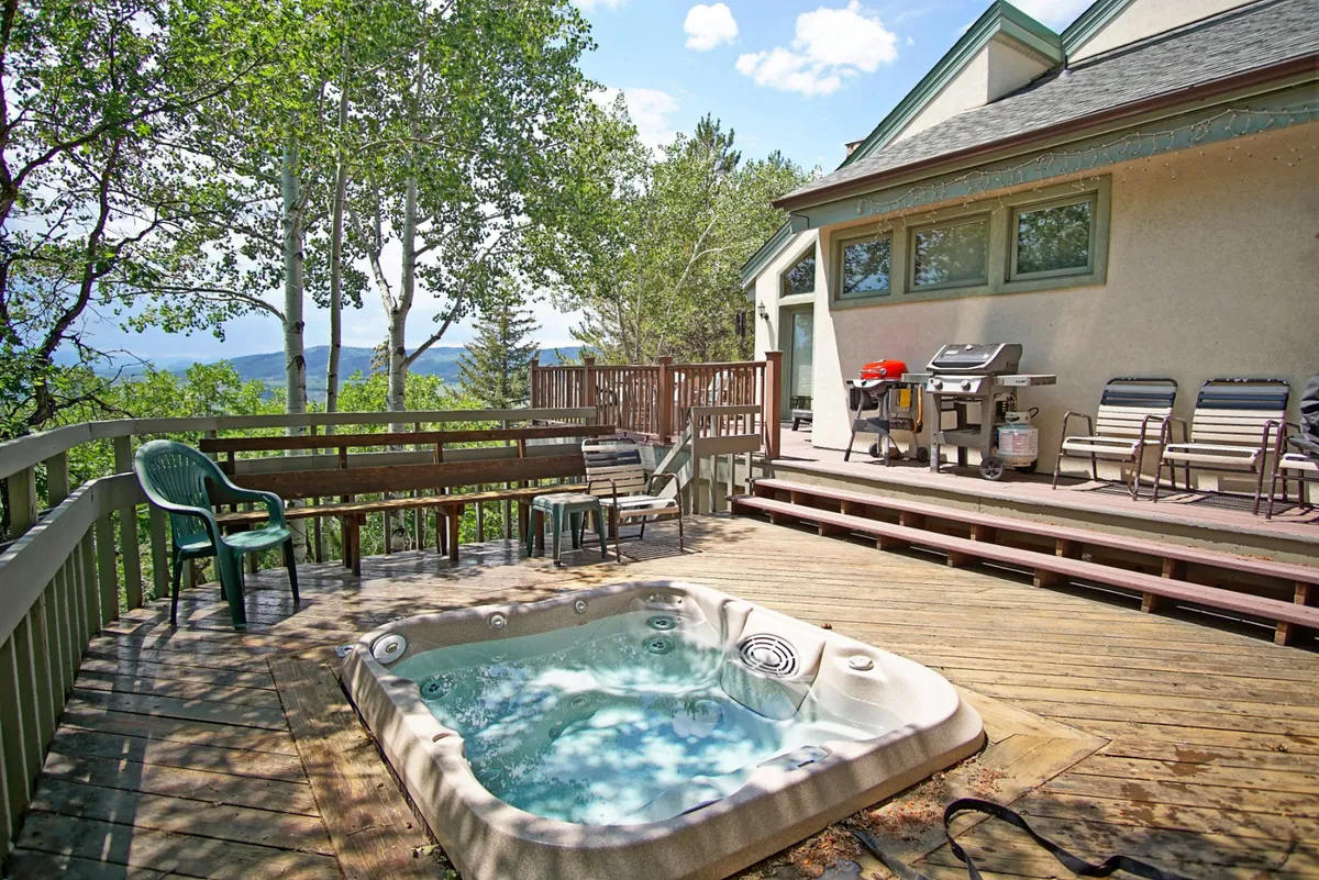 This image features a spacious deck nestled among mature trees, with a private hot tub, comfortable lounge seating, a grill for barbecues, and tranquil forest surroundings. The setting captures the seamless indoor-outdoor connection and offers glimpses of the natural landscape.