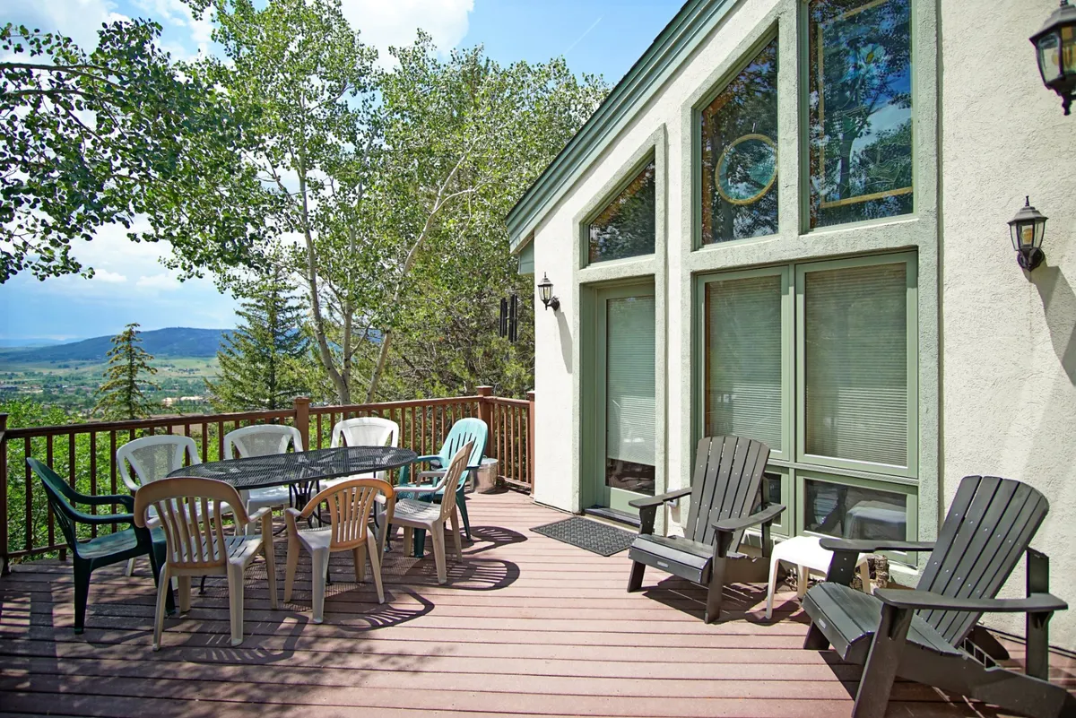 An outdoor entertaining space with a dining table and Adirondack chairs on a wooden deck, set against a backdrop of lush trees and the house’s exterior. The atmosphere is cheerful, ideal for group gatherings or quiet mornings with coffee.