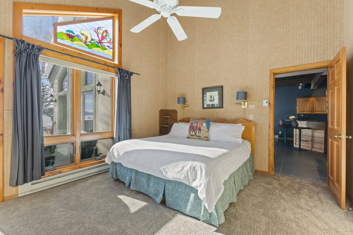 A cozy bedroom with a comfortable bed, curtains framing a stained glass window, and tasteful wall decor. The space feels intimate and inviting, perfect for a restful night's sleep or quiet reading.