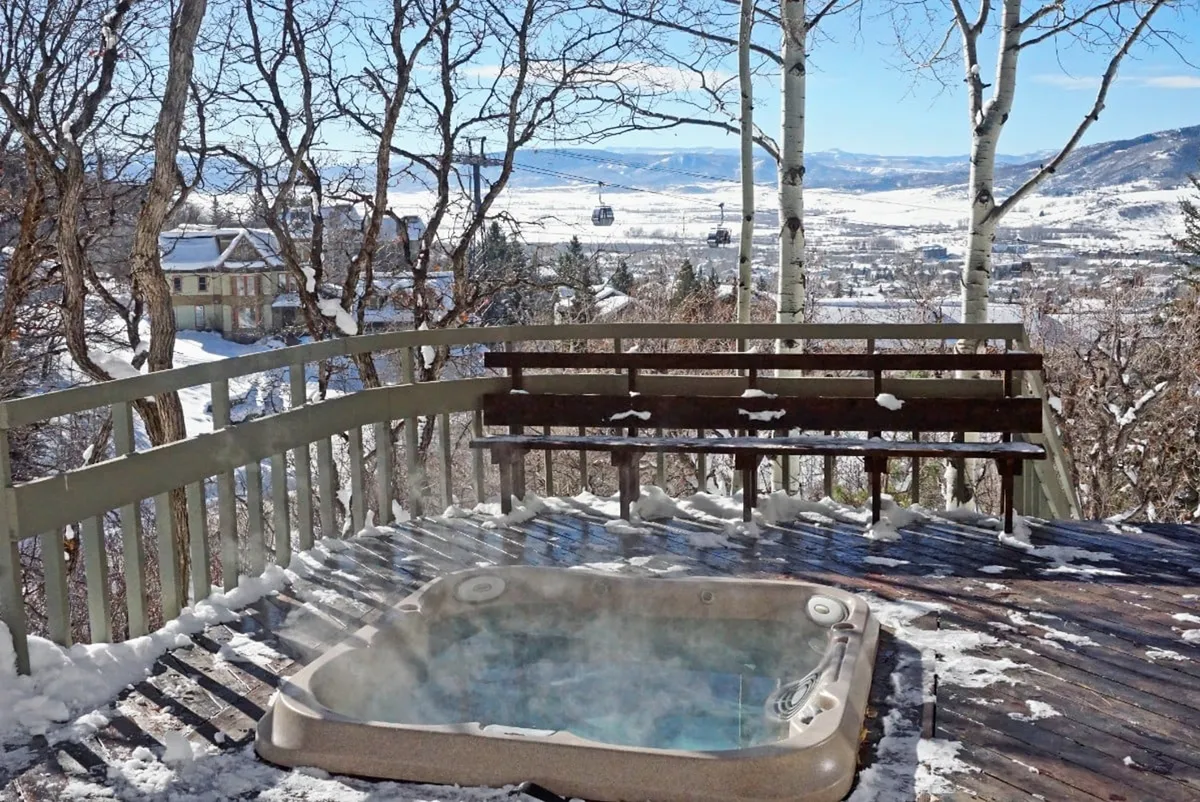 An inviting outdoor hot tub sits on a deck overlooking a snow-covered landscape, with nearby mountains and the Steamboat gondola visible in the background. The hot tub setting is tranquil, hinting at après-ski relaxation with panoramic winter views.