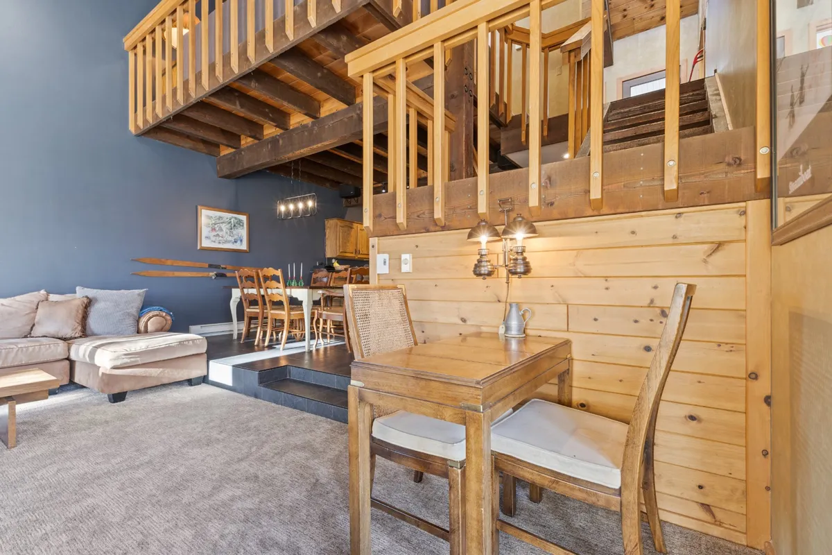 A cozy, sunlit interior featuring a wooden dining table, a comfortable sofa with cushions, a staircase leading to the upper level, and a wooden railing. The space feels welcoming and inviting, perfect for relaxing, dining with family, or working remotely.